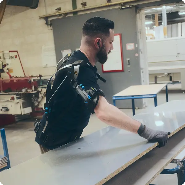 Mitarbeiter von Keller & Wehbrink Möbelteile GmbH mit einem Exoskelett bewegt Holzplatten für die Sockelleistenproduktion.